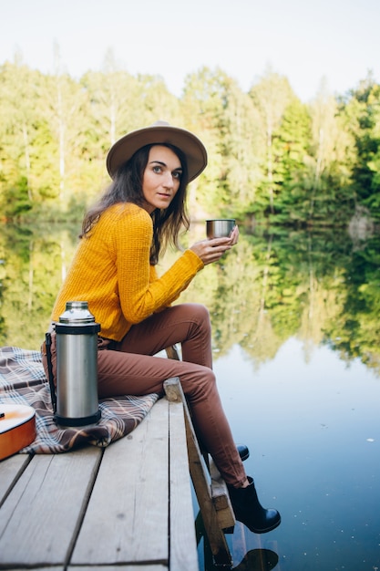 La giovane donna si siede su un ponte su un lago con un paesaggio autunnale e beve tè caldo da un thermos. Tonificante.