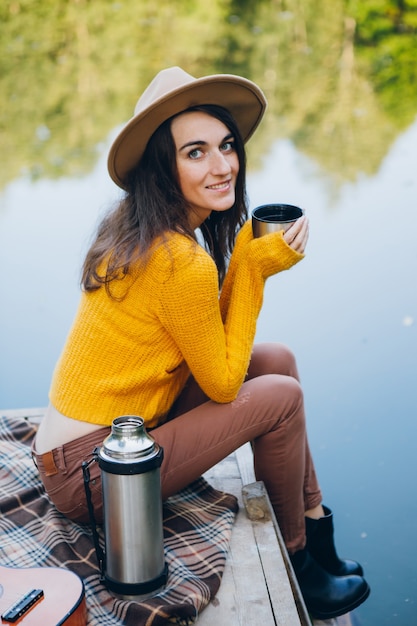 La giovane donna si siede su un ponte su un lago con un paesaggio autunnale e beve tè caldo da un thermos. Tonificante.