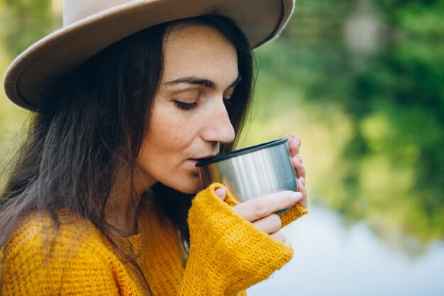 La giovane donna si siede su un ponte su un lago con un paesaggio autunnale e beve tè caldo da un thermos. Tonificante.