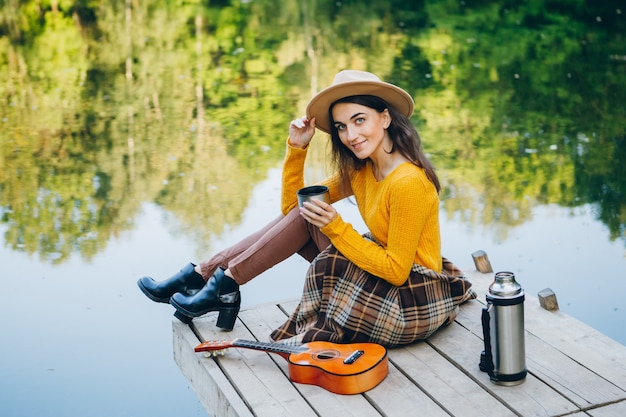 La giovane donna si siede su un ponte su un lago con un paesaggio autunnale e beve tè caldo da un thermos. Tonificante.