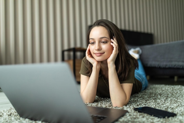 La giovane donna si siede sul tappeto piegato usa il laptop per realizzare un progetto di gestione del lavoro nella stanza al chiuso