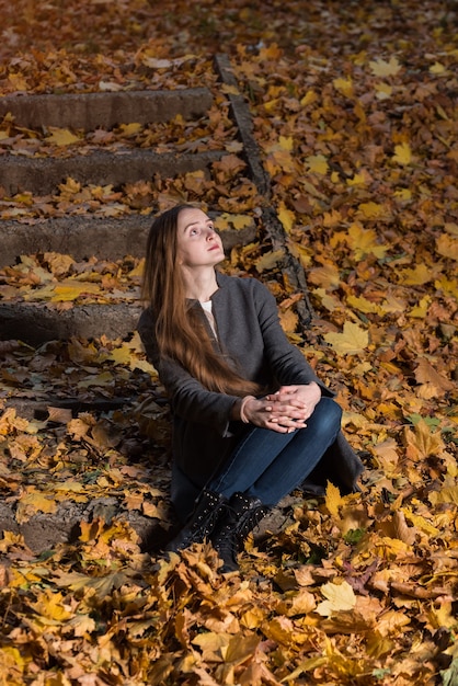 La giovane donna si siede sulle foglie cadute in autunno Park. Cornice verticale.