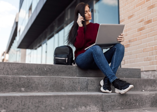 La giovane donna si siede sulle scale urbane e lavora con il computer portatile sulla strada