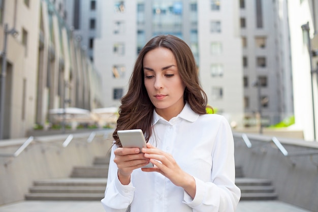 La giovane donna sicura di affari felice sta usando il suo smartphone mentre cammina all'aperto in una città urbana