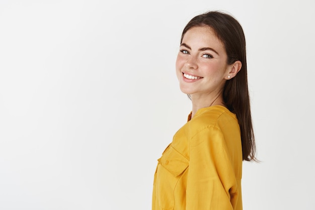 La giovane donna sicura di sé gira la testa davanti, sembra felice e sorridente, in piedi sul muro bianco con una camicetta gialla