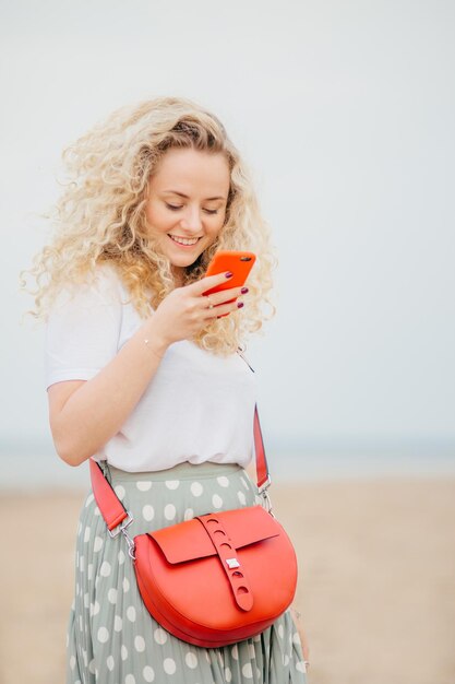 La giovane donna soddisfatta con i capelli ricci folti tiene lo smartphone utilizza la connessione Internet gratuita per inviare messaggi di testo su sfondo sfocato del paesaggio marino Concetto di persone e attività ricreative