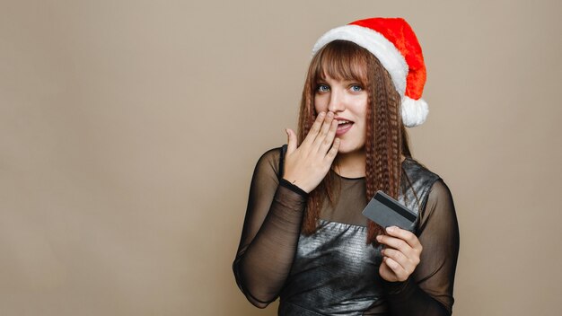 La giovane donna sorpresa in cappello del Babbo Natale tiene una carta di credito o di debito. copia spazio. Banner web.