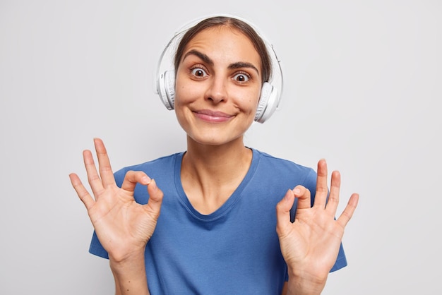 La giovane donna sorpresa soddisfatta mostra un gesto ok con entrambe le mani ti assicura che tutto è ok indossa cuffie stereo wireless sulle orecchie vestite con una maglietta blu casual isolata sopra il muro bianco