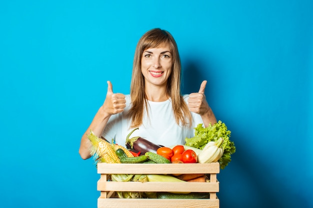 La giovane donna sorride e tiene una scatola con gli ortaggi freschi sul blu. Buon concetto di vendemmia, prodotto naturale