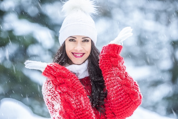 La giovane donna sorride nella foresta innevata indossando capispalla di moda come maglione rosso, guanti bianchi e cappello con pompon.