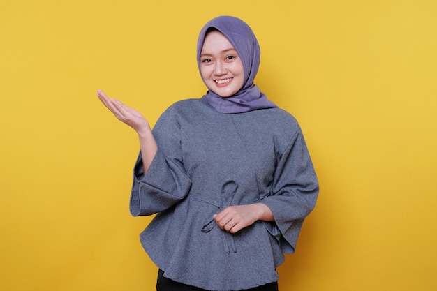 La giovane donna sorridente che indossa l'hijab mostra qualcosa e tiene qualcosa con i palmi isolati su uno sfondo giallo chiaro