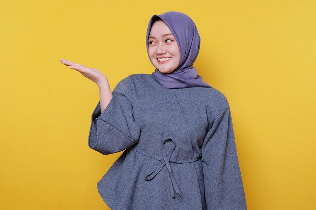 La giovane donna sorridente che indossa l'hijab mostra qualcosa e tiene qualcosa con i palmi isolati su uno sfondo giallo chiaro