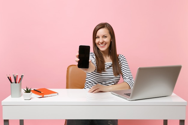 La giovane donna sorridente che mostra il telefono cellulare con lo schermo vuoto vuoto lavora alla scrivania bianca con un computer portatile contemporaneo