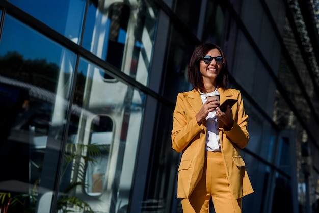 La giovane donna sorridente di affari in occhiali da sole e vestito giallo sta vicino al centro dell'ufficio, beve il caffè e utilizza lo smartphone moderno.