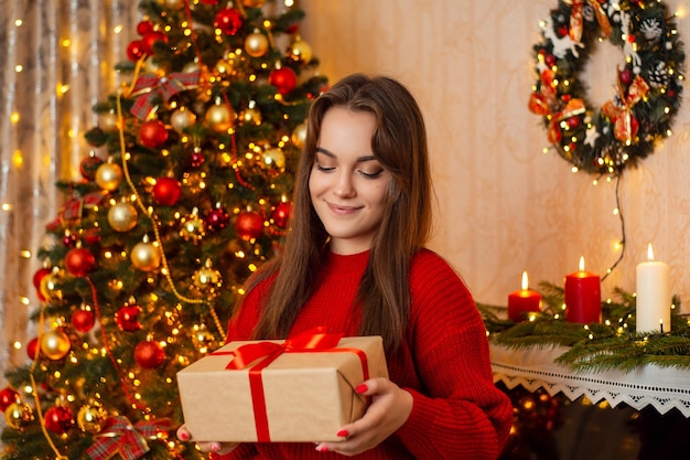 La giovane donna sorridente sceglie una scatola con un regalo di Natale nelle sue mani rilassandosi a casa godendosi la decorazione magica festiva di Natale nella stanza
