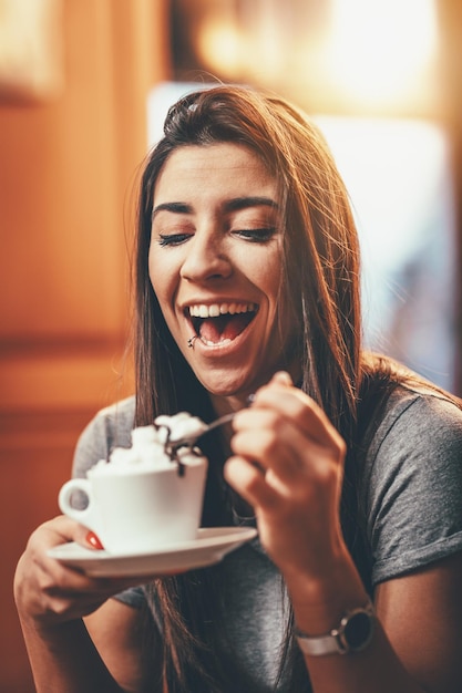 La giovane donna sorridente sta bevendo il caffè con la panna montata mentre si siede nella caffetteria