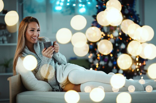 La giovane donna sorridente sveglia è seduta sul letto con in mano una tazza di caffè, circondata da luci a bolle di Natale.