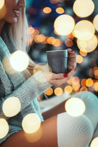 La giovane donna sorridente sveglia è seduta sul letto e tiene una tazza di caffè o tè, circondata da luci di Natale.