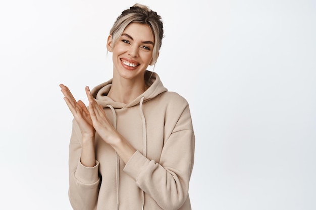 La giovane donna sorridente ti applaude guardando felice e battendo le mani congratulandosi con qualcuno in piedi su sfondo bianco