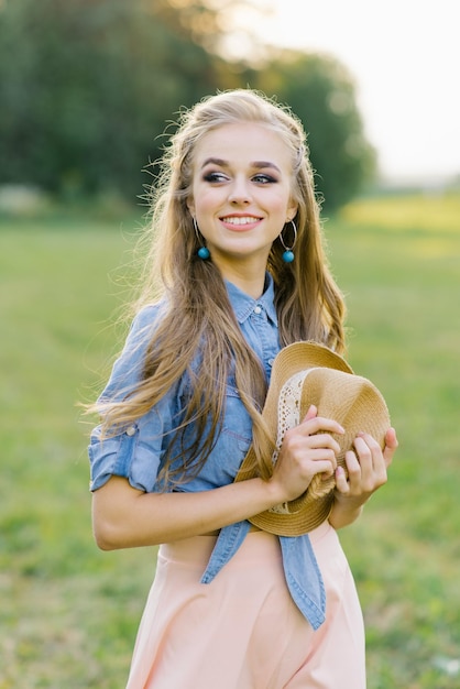 La giovane donna sorridente tiene un cappello in mano mentre cammina in estate all'aperto fuori città in vacanza