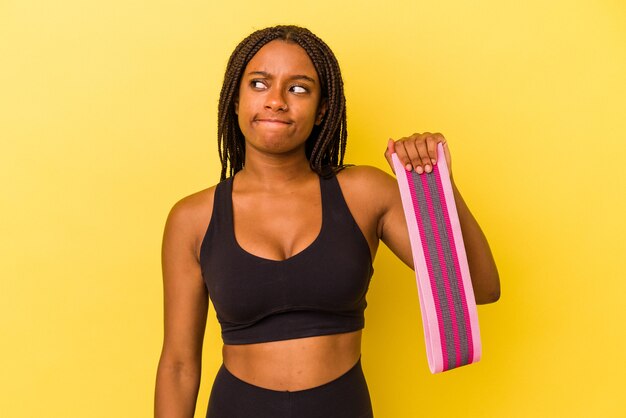 La giovane donna sportiva afroamericana che tiene un elastico isolato su sfondo giallo confuso, si sente dubbiosa e insicura.