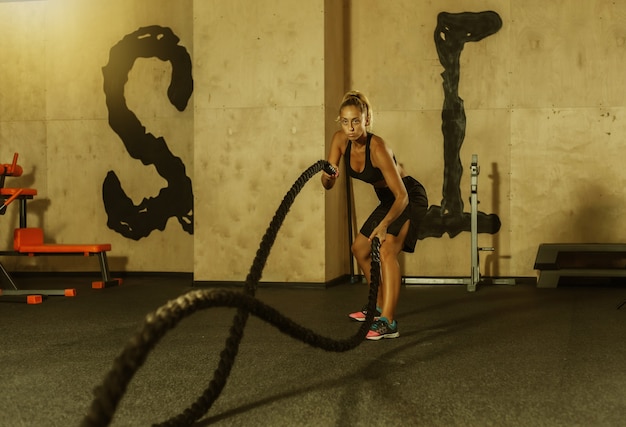 La giovane donna sportiva in abiti sportivi si sta allenando con la corda da battaglia in palestra. Allenamento funzionale. Concetto di stile di vita sano