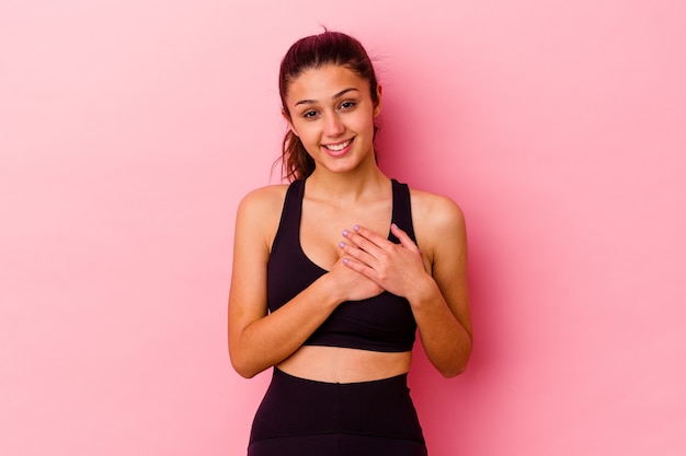 La giovane donna sportiva isolata sul muro rosa ha un'espressione amichevole, premendo il palmo sul petto