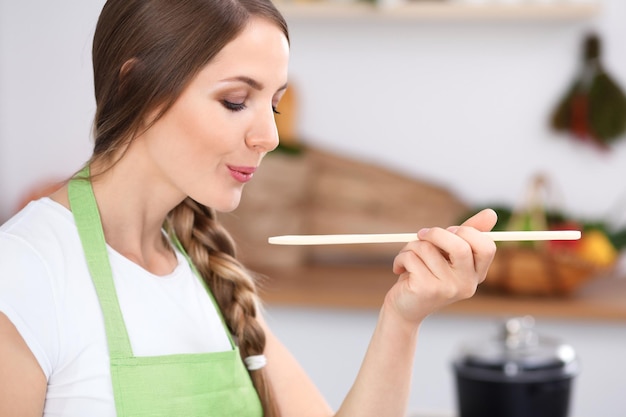 La giovane donna sta cucinando in una cucina La casalinga sta gustando la zuppa con un cucchiaio di legno