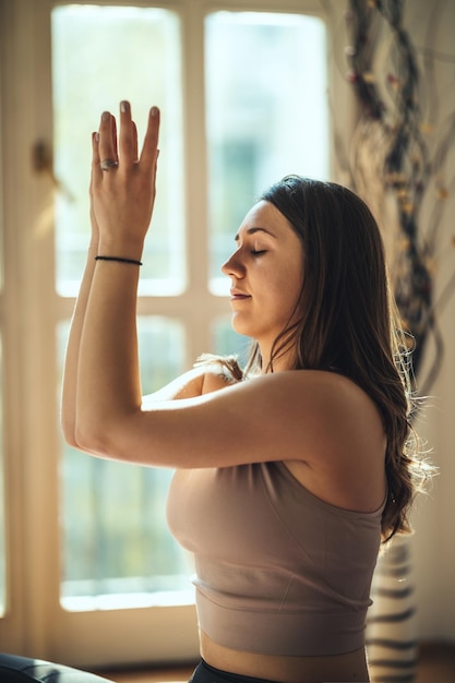 La giovane donna sta facendo meditazione yoga durante la pandemia di coronavirus nel soggiorno di casa. Sta meditando al sole del mattino.
