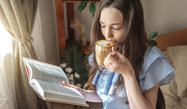 La giovane donna sta leggendo un libro e sta bevendo un caffè seduto vicino alla finestra Concetto di divertimento mattutino tempo libero piacevole passatempo e relax