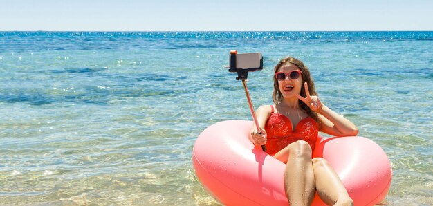 La giovane donna sta posando su un anello gonfiabile rosa e sta prendendo selfie con un monopiede in mare Riposo in spiaggia