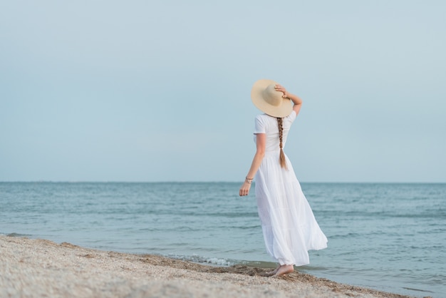 La giovane donna sta sulla spiaggia sabbiosa contro il mare e la tenuta con il cappello di paglia della mano