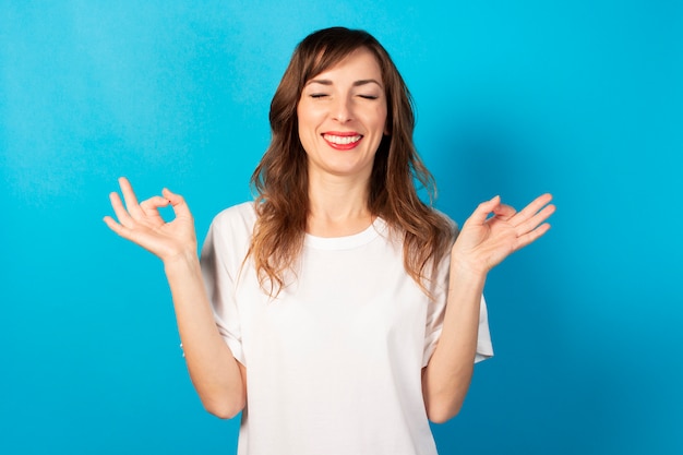 La giovane donna sveglia con gli occhi chiusi e una maglietta bianca fa un gesto con le sue mani yoga, concentrazione, meditazione sul blu isolato. Concetto di meditazione, sogni, pianificazione, buon umore