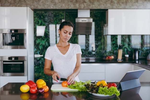 La giovane donna taglia le verdure in cucina con un coltello e un laptop sul tavolo