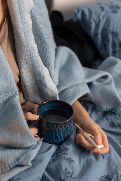 La giovane donna tiene in mano una tazza con una bevanda calda e un termometro. Ragazza affetta da malattia stagionale. Femmina che riposa a letto, recupero dopo il disturbo. Concetto di influenza. Sintomi di raffreddore e influenza.