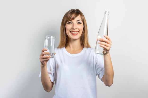 La giovane donna tiene una bottiglia e un bicchiere con acqua limpida su uno sfondo chiaro