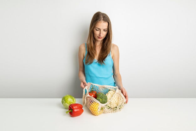 La giovane donna tira fuori le verdure dalla borsa sul tavolo su uno sfondo bianco
