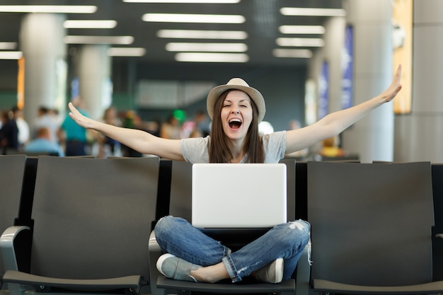 La giovane donna turistica divertente del viaggiatore con il computer portatile si siede con le gambe incrociate che allargano le mani come in volo, aspettando nella hall dell'aeroporto