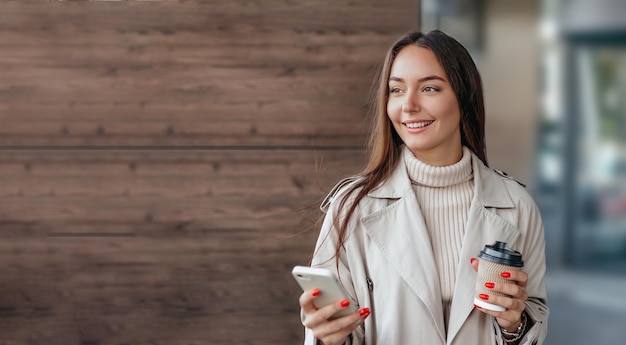 La giovane donna usa un telefono cellulare sorride e distoglie lo sguardo sullo sfondo di un edificio per uffici sulla strada