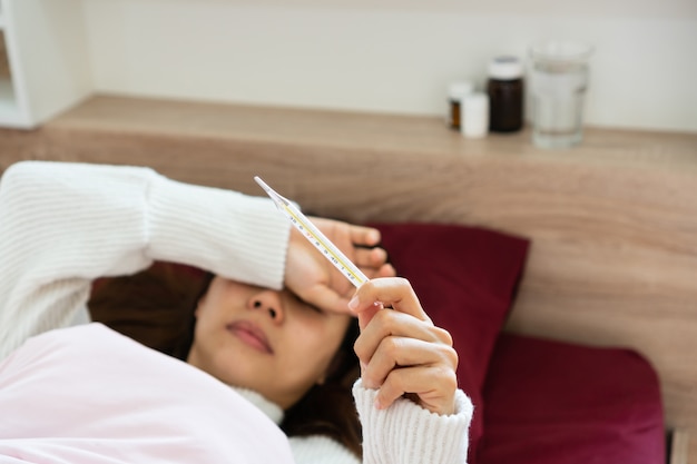 La giovane donna vaga soffre di febbre malata a letto sta tenendo in mano il termometro mentre si utilizza il tocco della mano sulla sua fronte. egli concetto di malattia e malattia