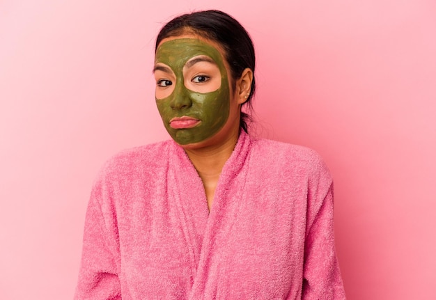 La giovane donna venezuelana che indossa un accappatoio e una maschera facciale isolata su sfondo rosa scrolla le spalle e apre gli occhi confusi