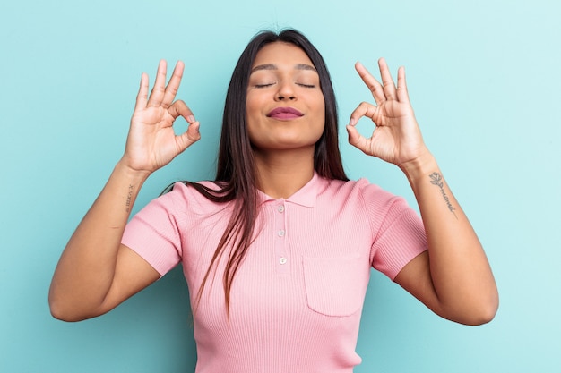 La giovane donna venezuelana isolata su sfondo blu si rilassa dopo una dura giornata di lavoro, sta eseguendo yoga.