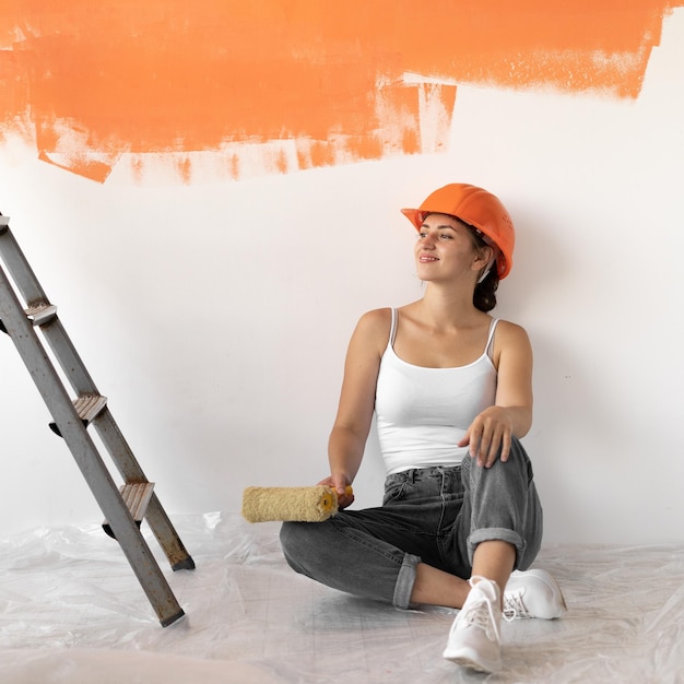 La giovane donna vestita con un elmetto arancione si siede sul pavimento vicino al muro. Concetto di ristrutturazione della casa.