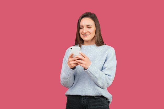 La giovane femmina positiva sta tenendo il suo telefono e sta digitando qualcosa Studio girato su sfondo rosa