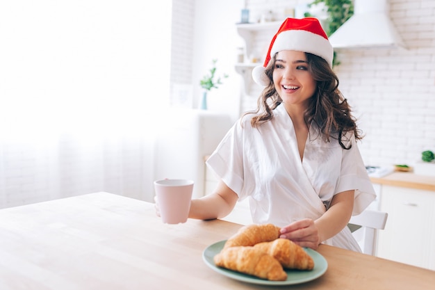 La giovane governante spensierata attraente fa colazione in mattina in cucina. Indossa un cappello rosso di Natale. Festeggiamo il nuovo anno. Umore festivo. Donna felice positiva allegra. Daylight.