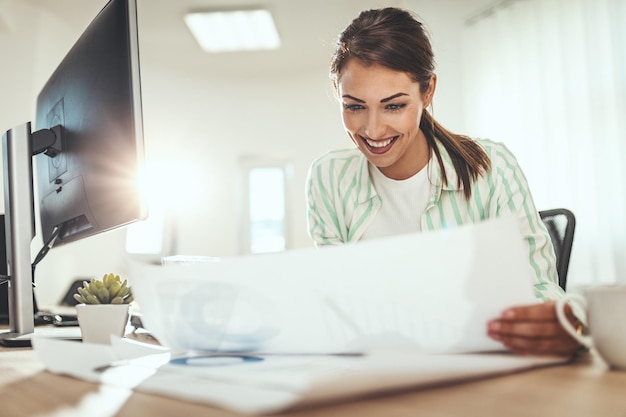 La giovane imprenditrice sorridente di successo sta lavorando in ufficio, analizzando il progetto e bevendo caffè.