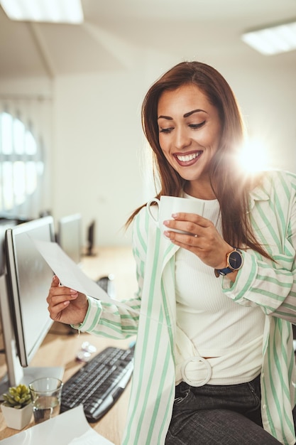 La giovane imprenditrice sorridente di successo sta lavorando in ufficio, analizzando il progetto e bevendo caffè.