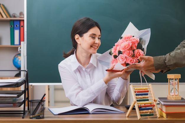 la giovane insegnante sorridente ha ricevuto un mazzo di fiori seduto alla scrivania con gli strumenti della scuola in classe