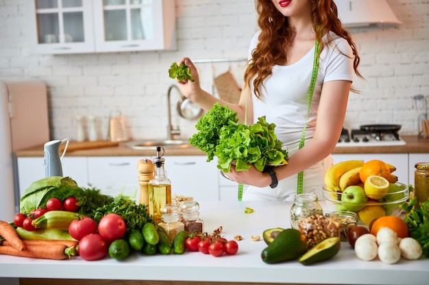 La giovane lattuga felice della tenuta della donna lascia per produrre l'insalata nella bella cucina con gli ingredienti freschi verdi all'interno. Cibo sano e concetto di dieta. Perdere peso