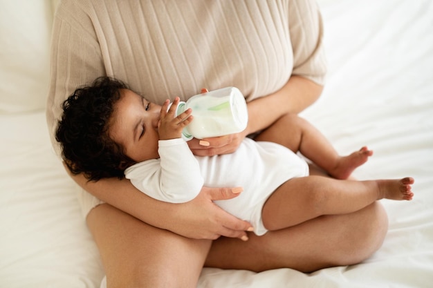 La giovane madre afroamericana si siede sul letto che alimenta il bambino piccolo con la bottiglia che si rilassa insieme nella camera da letto